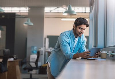 Young businessman leaning against stand up table in a bright modern office working on a digital tablet