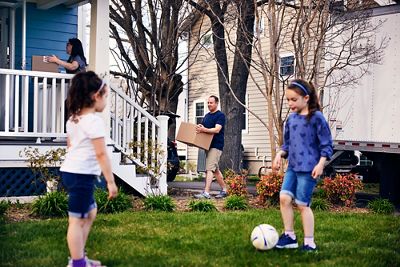 Parents bringing in moving boxes from moving van as daughters play in the yard